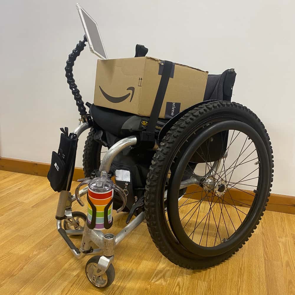 A manual wheelchair has a box on the seat and lots of great accessories attached to it, such as a phone mount, a cup holder and a lap strap to hold the box in place.